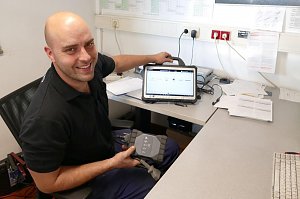 Ein freundlicher Mitarbeiter im schwarzen Polohemd sitzt an einem Schreibtisch in einem Büro. Er hält ein Diagnosegerät in der Hand und zeigt lächelnd auf ein Tablet mit einer Fahrzeug-Software.