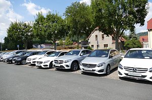 Reihe von Mercedes-Benz Gebrauchtwagen in verschiedenen Farben, geparkt auf einem Autohaus-Außenplatz. Im Hintergrund stehen Bäume, ein Wohnhaus mit rotem Dach und weitere Häuser, blauer Himmel mit einigen Wolken.