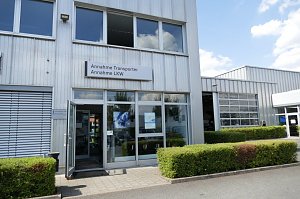 Eingangsbereich eines Autohauses mit Glasfront. Über der Tür hängt ein Schild mit der Aufschrift ‚Annahme Transporter, Annahme LKW‘. Rechts ist eine Werkstatthalle mit großen Toren sichtbar, davor gepflegte Hecken und ein gepflasterter Weg.
