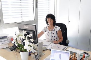 Frau mit dunklen Haaren sitzt an einem Schreibtisch in einem hellen Büro. Sie arbeitet am Computer und schaut in die Kamera. Auf dem Schreibtisch stehen eine Orchidee, Akten und Büromaterial.