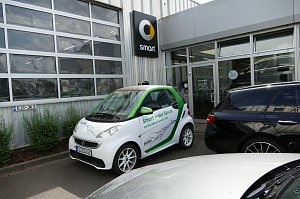 Wei&szlig;-gr&uuml;ner Smart mit der Aufschrift �Smart unter Strom und die Zukunft geh&ouml;rt DIR!�, geparkt vor einem Autohaus. Auf dem Auto ist ein Blitzmotiv abgebildet. Im Hintergrund ist das Smart-Logo an der Geb&auml;udewand zu sehen.