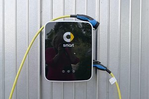 Ladestation mit Smart-Logo an einer silberfarbenen Metallwand. Die Station hat ein schwarzes Frontpanel mit gelbem Kabel und blau-schwarzem Stecker, der an der Seite h&auml;ngt.