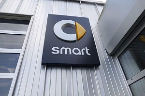 Nahaufnahme eines schwarzen Schildes mit dem Smart-Logo an einer silberfarbenen Metallfassade. Das Logo besteht aus einem stilisierten grauen �C� mit einem gelben, pfeilf&ouml;rmigen Element, darunter steht der Schriftzug �smart� in wei&szlig;en Buchstaben.