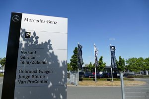 Gro&szlig;es wei&szlig;es Mercedes-Benz Hinweisschild mit Smart-Logo, Beschriftung f&uuml;r Verkauf, Service, Teile/Zubeh&ouml;r, Gebrauchtwagen, Junge Sterne und Van ProCenter; im Hintergrund wehen mehrere Mercedes- und Smart-Flaggen, B&auml;ume und geparkte Autos unter blauem Himmel.