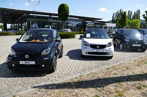 Reihe von Smart-Autos (schwarz und wei&szlig;) auf dem Parkplatz vor einem Mercedes-Benz Autohaus mit Peter-Logo in Northeim, im Hintergrund moderner Glasbau, gepflegte B&uuml;sche und B&auml;ume unter blauem Himmel.