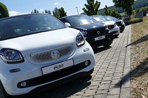 Nahaufnahme einer Reihe von Smart-Autos in Wei&szlig; und Schwarz, schr&auml;g geparkt auf einem gepflasterten Parkplatz vor dem Autohaus Peter in Northeim, sonniger Tag mit B&auml;umen und H&uuml;geln im Hintergrund.