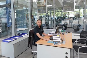 Mitarbeiter in schwarzem Mercedes-Benz Polohemd sitzt an einem modernen Holzschreibtisch im Autohaus, mit Computer, Dokumenten und St&uuml;hlen f&uuml;r Kunden, Glasw&auml;nde mit Werbepostern und Fahrzeugen im Hintergrund.