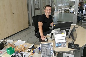 Junger, freundlich l&auml;chelnder Mitarbeiter sitzt an einem Schreibtisch im Autohaus. Er tr&auml;gt ein schwarzes Poloshirt mit Mercedes-Logo. Vor ihm stehen ein Computer, viele Unterlagen, Stempel, Prospekte und Schilder mit Service-Angeboten. Im Hintergrund Glast&uuml;ren und ein heller Empfangsbereich.