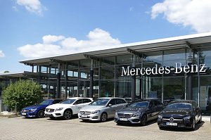 Mercedes-Benz Autohaus mit moderner Glasfassade und großem Mercedes-Benz-Schriftzug. Davor stehen fünf Mercedes-Benz Fahrzeuge in verschiedenen Farben (blau, weiß, silber, grau, schwarz) nebeneinander aufgereiht. Blauer Himmel mit weißen Wolken im Hintergrund.