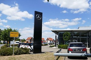 Mercedes-Autohaus mit großer Glasfassade und Mercedes-Stern aus Buschwerk. Links ein gelbes Ortsschild mit der Aufschrift ‚Petershütte Stadt Osterode am Harz‘. Mehrere geparkte Autos, blauer Himmel mit weißen Wolken.