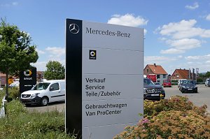 Großes Mercedes-Benz Schild auf einem Autohaus-Gelände. Auf dem Schild stehen die Angebote: Verkauf, Service, Teile/Zubehör, Gebrauchtwagen, Van ProCenter. Im Hintergrund mehrere Mercedes-Fahrzeuge, ein weiteres Smart-Schild und rote Hausdächer. Blauer Himmel mit einigen Wolken.