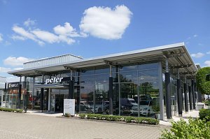 Modernes Mercedes-Benz Autohaus mit großer Glasfassade und der Aufschrift ‚peter‘ über dem Eingang. Links daneben ein Bereich mit Smart-Logo. Vor dem Gebäude gepflegte Grünflächen, blauer Himmel mit wenigen weißen Wolken.