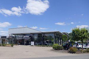 Glasfassade des Autohauses Peter mit Mercedes-Benz und Smart Logos. Vor dem Gebäude stehen mehrere Autos, im Vordergrund Bäume und Büsche. Der Himmel ist blau mit vereinzelten Wolken.