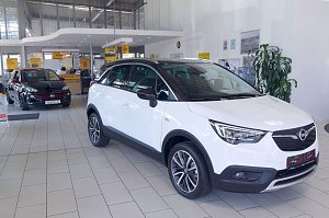 Im Innenraum eines Autohauses stehen zwei Opel-Fahrzeuge. Im Vordergrund ein wei&szlig;er Opel Crossland mit schwarzen Akzenten, im Hintergrund ein schwarzer Opel Adam mit rotem Dach. Der helle Ausstellungsraum ist modern gestaltet, mit gro&szlig;en Fenstern und einem Verkaufsbereich im Hintergrund. Der Boden ist hell gefliest. 