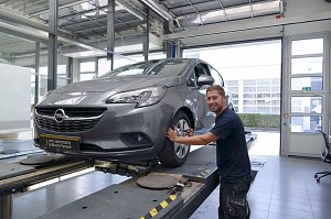 Ein grauer Opel steht auf der Hebeb&uuml;hne der Opel-Werkstatt. Ein junger Mechaniker l&auml;chelt in die Kamera, w&auml;hrend er mit einem Werkzeug am Vorderrad arbeitet. Im Hintergrund sind gro&szlig;e Fenster und ein Garagentor zu sehen, durch das Tageslicht hereinf&auml;llt. Auf dem Kennzeichen steht Opel Zertifizierte Gebrauchtwagen.