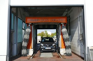 Smart-Auto f&auml;hrt in eine orangefarbene automatische Waschanlage mit rotierenden B&uuml;rsten, Schriftzug �Christ CAR WASH� &uuml;ber dem Eingang.