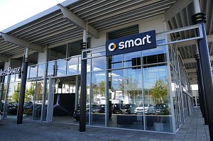 Glasfassade eines Autohauses mit Smart-Logo und Mercedes-Benz-Bereich, moderner Eingangsbereich mit gro&szlig;en Fenstern und metallischer Dachkonstruktion.