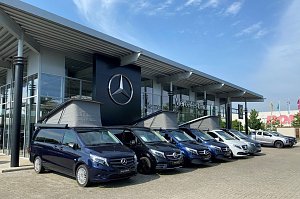 Au&szlig;enansicht des Mercedes-Benz Autohauses in Dessau. Vor dem modernen Glasgeb&auml;ude mit gro&szlig;em Mercedes-Stern-Logo stehen mehrere blaue und wei&szlig;e Mercedes-Vans in einer Reihe. Drei Vans haben ausgeklappte Aufstelld&auml;cher.