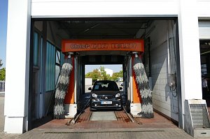 Ein grauer Smart f&auml;hrt gerade in eine Autowaschanlage mit orangefarbener Beschriftung �Christ CAR WASH�. Links und rechts h&auml;ngen graue B&uuml;rsten.