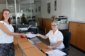 Eine junge Frau &uuml;berreicht einer Mitarbeiterin an einem Empfangsschalter freundlich ein Dokument. Die Mitarbeiterin sitzt hinter dem Schreibtisch und tr&auml;gt eine Brille. Beide Frauen l&auml;cheln. Im Hintergrund sieht man B&uuml;ror&auml;ume und Schr&auml;nke.