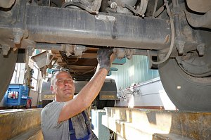 Ein Werkstattmitarbeiter steht unter einem gro&szlig;en LKW und arbeitet an dessen Unterseite. Er tr&auml;gt graues T-Shirt, schaut in die Kamera und l&auml;chelt. Im Hintergrund ist die Werkstatt mit Werkzeugen zu sehen.