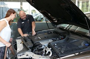 Ein Mercedes-Benz Servicemitarbeiter erkl&auml;rt einer jungen Frau den Motorraum eines Fahrzeugs. Die Motorhaube ist ge&ouml;ffnet, beide schauen gemeinsam in den Motor. Im Hintergrund helle Glasfronten des Autohauses.