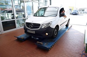 Ein wei&szlig;er Mercedes-Transporter mit Fahrer am Steuer steht in der Autohaus-Werkstatt auf einem blauen Pr&uuml;fstand, bereit zur Inspektion.