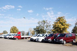 Reihe von Mercedes-Transportern und Vans, darunter ein roter Pritschenwagen, auf dem Autohaus-Parkplatz an einem sonnigen Tag mit blauem Himmel und B&auml;umen im Hintergrund.