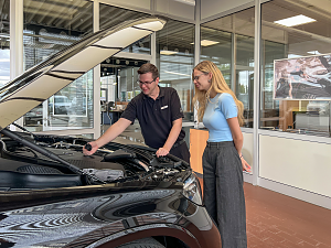 Ein Mitarbeiter im schwarzen Hemd mit Mercedes-Logo erkl&auml;rt einer jungen Kundin Details am Motorraum eines schwarzen Mercedes-Fahrzeugs.