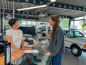 Mitarbeiterin am Empfangstresen im Mercedes-Benz Autohaus Bernburg &uuml;bergibt l&auml;chelnd Unterlagen an ein eine Kundin, moderner heller Showroom mit Computern, Prospekten und Autos im Hintergrund.