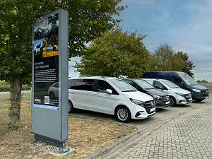 Reihe von verschiedenen Mercedes-Benz Vans auf dem Parkplatz des Autohauses, links ein gro&szlig;es Werbeschild, im Hintergrund B&auml;ume und blauer Himmel.