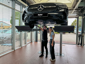 Die Dialogannahme des Autohauses mit gro&szlig;en Glasfronten. Ein schwarzes Mercedes-Fahrzeug befindet sich auf der angehobenen Hebeb&uuml;hne. Ein Mitarbeiter erkl&auml;rt einer Kundin Details am Unterboden des Autos.
