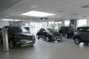 Heller, moderner Showroom des Opel-Autohauses mit mehreren ausgestellten Autos. Links vorne steht ein grauer Opel Mokka X, daneben ein kleinerer Opel Adam in der Mitte. Im Hintergrund sind weitere Fahrzeuge und Fahrr&auml;der zu sehen. Die gro&szlig;en Fensterfronten lassen viel Tageslicht herein, die Decke ist mit wei&szlig;en Paneelen und Einbaulampen ausgestattet. An der R&uuml;ckwand h&auml;ngen Farbmuster.