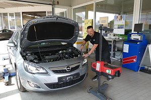 Ein Mechaniker &uuml;berpr&uuml;ft einen silbernen Opel mit offener Motorhaube in einer Werkstatt. Der Mechaniker steht freundlich l&auml;chelnd neben dem Fahrzeug und bedient ein Pr&uuml;fger&auml;t f&uuml;r die Scheinwerfer. Im Hintergrund sind Schreibtische, Poster und technische Ger&auml;te sichtbar.