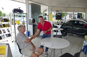 Zwei M&auml;nner in einem hellen Autohaus-Innenraum beim Handschlag. Der &auml;ltere Mann sitzt am Tisch, tr&auml;gt ein kariertes Hemd und Shorts, der j&uuml;ngere Mann in rotem Polo-Shirt steht und h&auml;lt Dokumente. Im Hintergrund sind ein schwarzes Auto, Tische, Prospektst&auml;nder und Regale mit Zubeh&ouml;r und Modellautos zu sehen. Gro&szlig;e Fenster lassen viel Tageslicht herein.