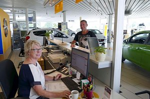 Empfangsbereich des Autohauses. Im Vordergrund sitzt eine freundliche Mitarbeiterin mit Brille am Schreibtisch vor einem Computer, sie schaut in die Kamera und l&auml;chelt. Im Hintergrund steht ein Kunde an der Theke und l&auml;chelt ebenfalls. Dahinter sind mehrere Autos im hellen, modernen Showroom zu sehen. Pflanzen und Dekoration sorgen f&uuml;r eine einladende Atmosph&auml;re.