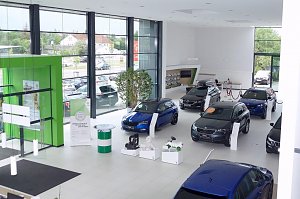 Heller �koda-Showroom mit gro&szlig;en Fensterfronten, mehreren ausgestellten Fahrzeugen, einem markanten gr&uuml;nen Eingangselement und Zubeh&ouml;r auf wei&szlig;en Podesten.
