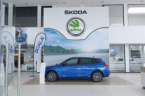 Innenansicht des �koda-Autohauses mit gro&szlig;em wei&szlig;en �koda-Logo an der Wand. In der Mitte steht ein blauer �koda Scala, flankiert von Werbefahnen und Postern.