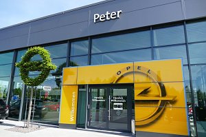 Eingang des Opel-Autohauses Peter in Nordhausen. &Uuml;ber der gl&auml;sernen Fassade steht der Schriftzug Peter. Vor dem Eingang befindet sich eine gro&szlig;e gelbe Wand mit Opel-Logo und Herzlich Willkommen-Schriftzug. Links davon steht eine kunstvoll geschnittene gr&uuml;ne Hecke in Form des Opel-Logos. Der Eingangsbereich wirkt modern und einladend.