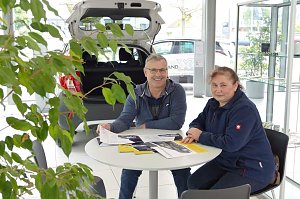 Ein Mann und eine Frau sitzen an einem Tisch im Autohaus, mit Brosch&uuml;ren und Prospekten vor sich, im Hintergrund ein ge&ouml;ffneter Kofferraum eines Autos und helle Glasfronten.