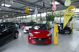 Innenansicht eines Autohauses mit mehreren ausgestellten Opel-Fahrzeugen, darunter ein roter Opel Adam im Vordergrund und ein schwarzer Opel Corsa im Hintergrund; gelbe Opel-Werbeflaggen und bunte Dekoelemente h&auml;ngen von der Decke.