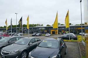 Opel-Autohaus Peter in G&ouml;ttingen mit moderner Glasfassade, mehreren gelben Opel-Flaggen im Vordergrund und zahlreichen geparkten Fahrzeugen auf dem Au&szlig;engel&auml;nde.