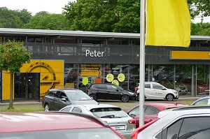 Ansicht des Opel Autohauses Peter mit gro&szlig;en Glasfassaden, gelbem Opel-Logo &uuml;ber dem Eingang und mehreren geparkten Autos im Vordergrund; gelbe Fahne und B&auml;ume im Hintergrund.