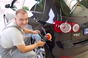 Pierre Heyer poliert die Heckseite eines schwarzen VW Golf Plus. Er tr&auml;gt graue Arbeitskleidung, h&auml;lt eine Poliermaschine in der Hand und schaut l&auml;chelnd in die Kamera. Das Fahrzeug gl&auml;nzt stark, Spiegelungen sind deutlich zu sehen.