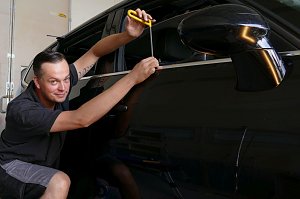 Kurt Kuschmierz arbeitet l&auml;chelnd an der T&uuml;r eines schwarzen Autos. Er benutzt ein Spezialwerkzeug mit gelbem Griff, um eine Delle vorsichtig auszubeulen. Er kniet neben dem Fahrzeug und zeigt dabei Feingef&uuml;hl und Pr&auml;zision. Werkstattumgebung im Hintergrund.