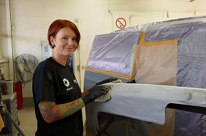 Katja R&ouml;pcke f&uuml;hrt Schleifarbeiten an einem grundierten Karosserieteil durch. Sie tr&auml;gt schwarze Handschuhe und ein schwarzes T-Shirt mit Firmenlogo, hat rote Haare und sichtbare Tattoos am Arm. Im Hintergrund ein abgeklebtes Fahrzeug in einer Werkstatt.