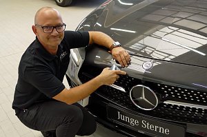 Dirk Morgenthal, Leiter des Lackierzentrums, kniet neben einem schwarzen Mercedes mit dem Schild �Junge Sterne�. Er tr&auml;gt ein schwarzes Polohemd mit Aufschrift �Peter� und und h&auml;lt ein Farbmessger&auml;t an die Motorhaube, um den exakten Farbton zu bestimmen. Heller Werkstattboden, reflektierende Autolackierung.