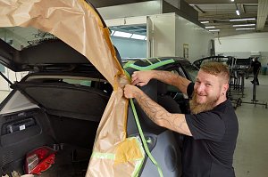 Frank Frank l&auml;chelt, w&auml;hrend er ein Fahrzeug f&uuml;r Lackierarbeiten mit braunem Abdeckpapier abklebt. Das Auto steht mit ge&ouml;ffneter Heckklappe in einer Werkstatt. Im Hintergrund sind weitere Fahrzeuge und Arbeitsbereiche zu sehen.