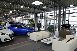 Heller, moderner Showroom eines Autohauses mit mehreren Peugeot-Fahrzeugen, unter anderem einem blauen Kleinwagen. Im Vordergrund eine Warteecke mit wei&szlig;en Sesseln und Couchtisch, im Hintergrund drei M&auml;nner im Gespr&auml;ch.