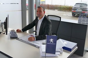 Berater in einem Peugeot-Autohaus sitzt an einem Schreibtisch mit Computer, Unterlagen und einer kleinen blauen Peugeot-Fahne, im Hintergrund Autos vor dem Fenster sichtbar.